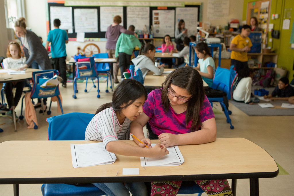 In a busy fourth-grade classroom, two students work together on a document while other small groups engage in lively discussion.
