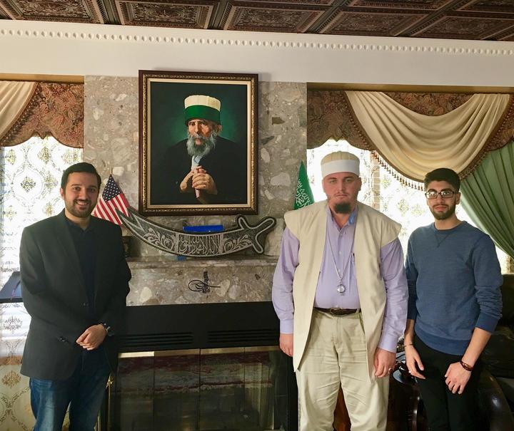Three men standing, relaxed, one is smiling, in front of picture of bearded Arab man in front of fireplace