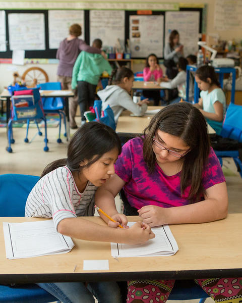 In a busy fourth-grade classroom, two students work together on a document while other small groups engage in lively discussion.